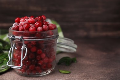 Photo of Frozen red cranberries in glass jar and green leaves on brown textured table, closeup. Space for text