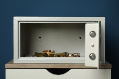 Open steel safe with gold bars and jewelry on wooden table against blue background