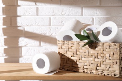 Photo of Toilet paper rolls in wicker basket and green leaves on wooden shelf against white brick wall