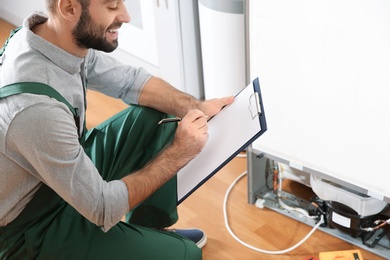 Photo of Male technician with clipboard examining refrigerator indoors
