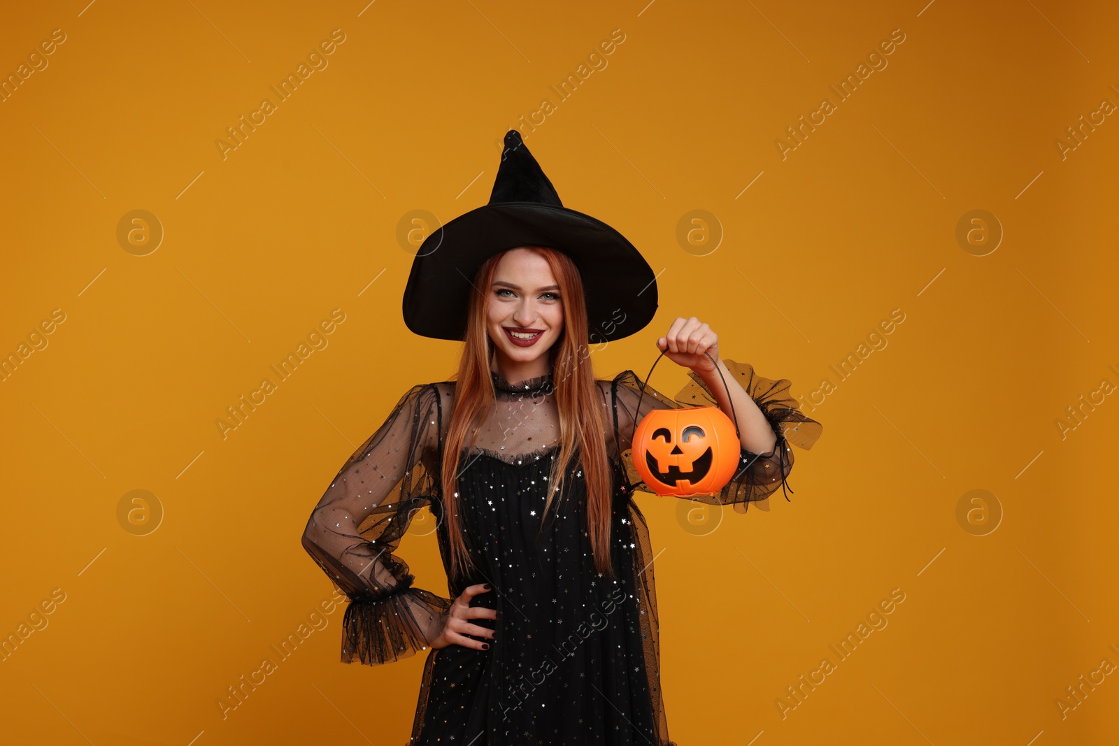 Photo of Happy young woman in scary witch costume with pumpkin bucket on orange background. Halloween celebration