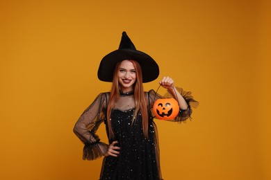 Happy young woman in scary witch costume with pumpkin bucket on orange background. Halloween celebration