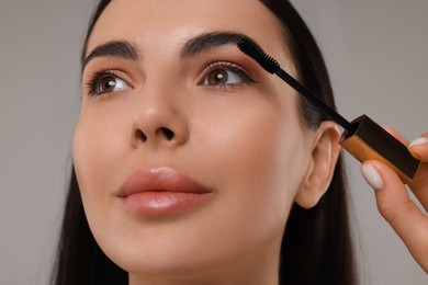 Beautiful young woman applying mascara on grey background, closeup