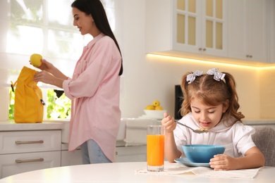 Photo of Little girl having breakfast while mother helping her get ready for school in kitchen