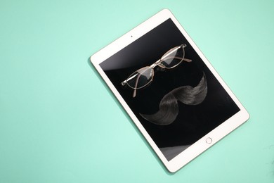 Photo of Artificial moustache, tablet and glasses on turquoise background, top view. Space for text