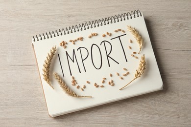 Notebook with word import and ears of wheat on wooden table, top view