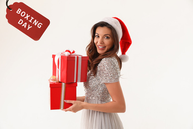 Image of Beautiful woman wearing Santa hat holding Christmas gifts and tag with text Boxing day on white background