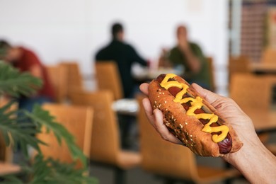 Photo of Woman holding fresh tasty hot dog with mustard in cafe, closeup. Space for text