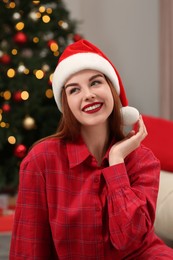 Photo of Beautiful young woman in Santa hat near Christmas tree at home