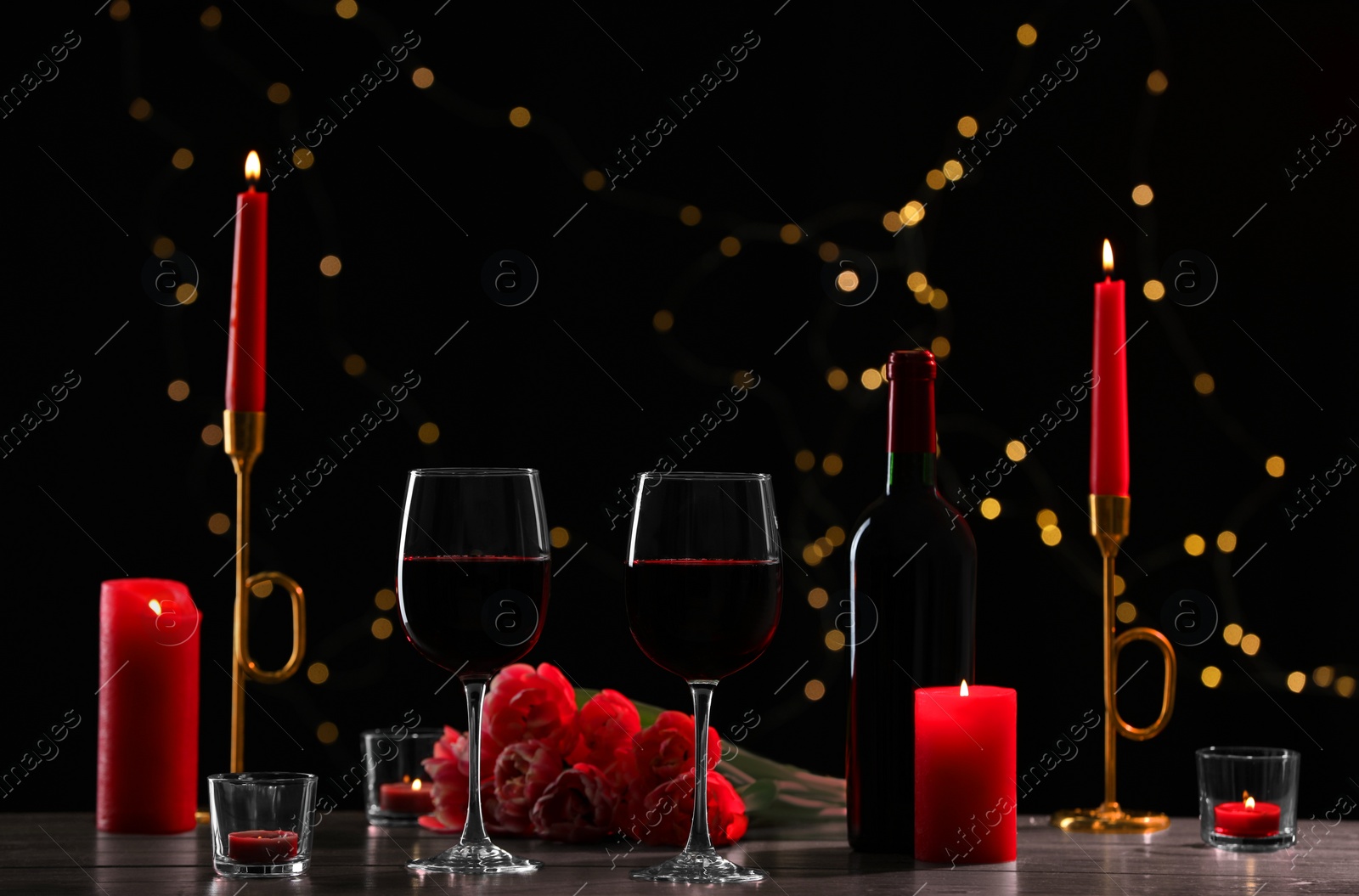 Photo of Glasses of red wine, tulip flowers and burning candles on black background against blurred lights. Romantic atmosphere