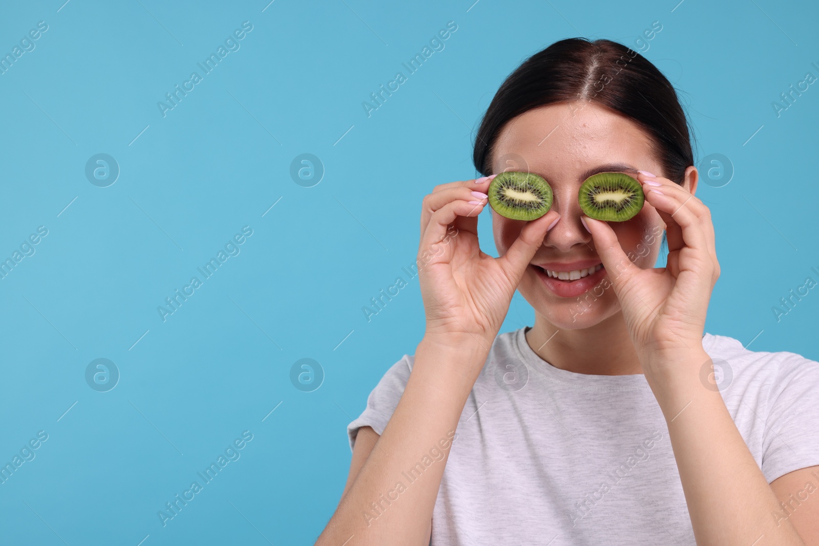 Photo of Young woman holding halves of kiwi near her eyes on light blue background, space for text