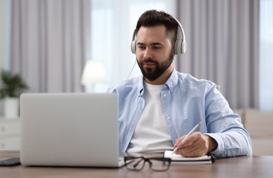 Photo of Young man in headphones watching webinar at table in room