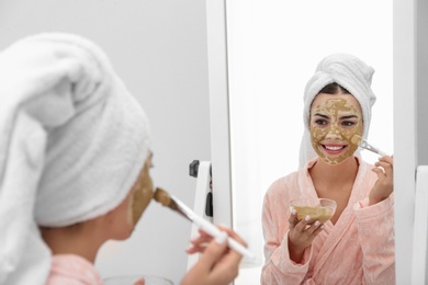 Photo of Young woman applying clay mask on her face near mirror in bathroom. Skin care