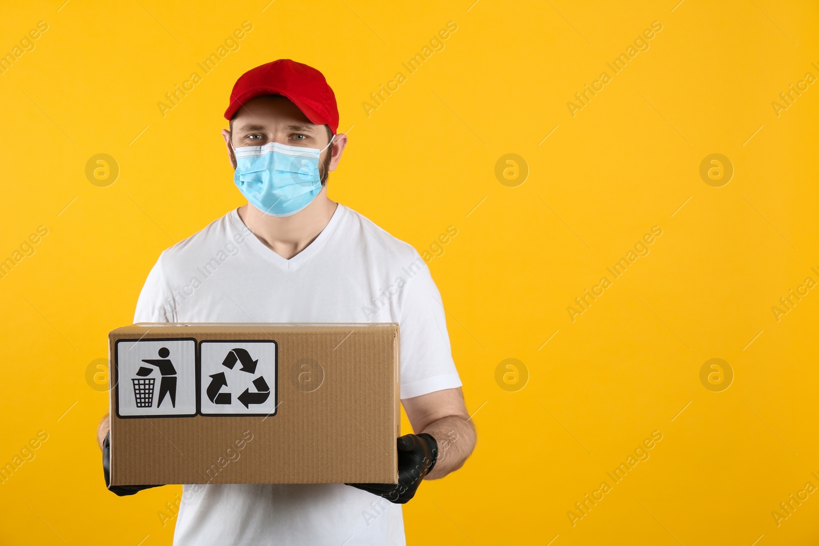 Photo of Courier in mask holding cardboard box with different packaging symbols on yellow background, space for text. Parcel delivery