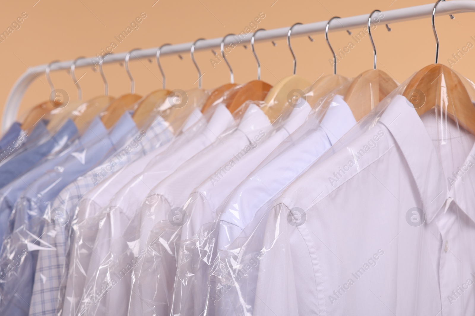 Photo of Dry-cleaning service. Many different clothes in plastic bags hanging on rack against beige background, closeup