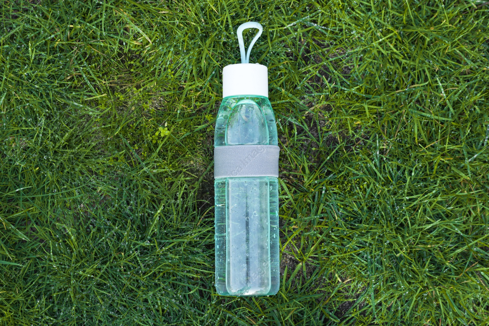 Photo of Glass bottle of fresh water on green grass outdoors, top view