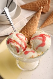 Delicious scoops of vanilla ice cream with wafer cone and strawberry topping in glass dessert bowl on table, closeup