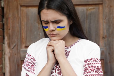 Sad young woman with drawings of Ukrainian flag on face near wooden door