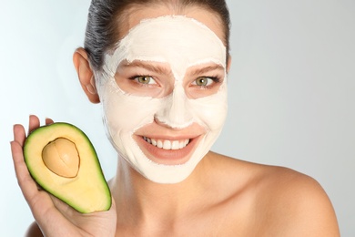 Photo of Beautiful woman holding avocado near her face with clay mask against grey background, closeup