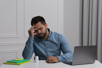 Photo of Depressed man at white table with antidepressants, laptop and stationery indoors