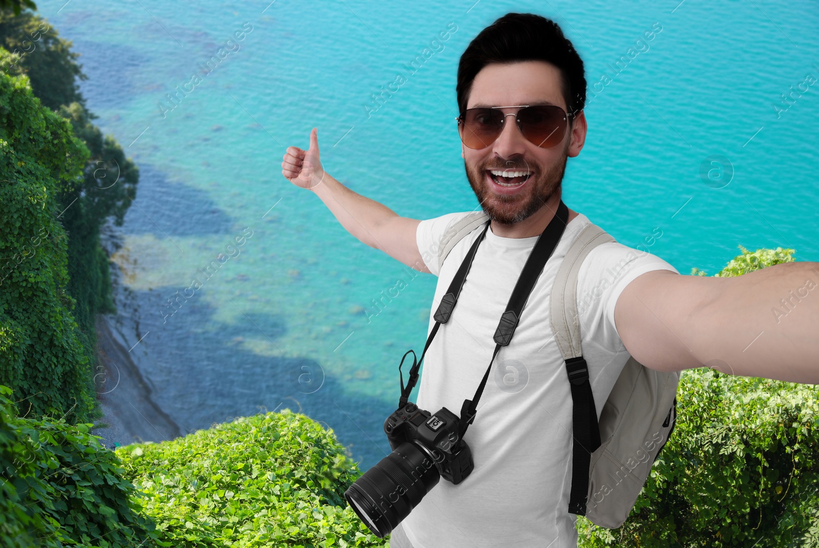 Image of Smiling man with camera taking selfie and showing thumbs up on green hill near sea