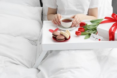 Photo of Tasty breakfast served in bed. Woman with tea, macarons, gift box, flowers and I Love You card at home, closeup