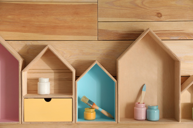 Photo of House shaped shelves with jars of paints and brushes on wooden wall. Interior elements