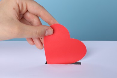 Photo of Woman putting red heart into slot of donation box against light blue background, closeup