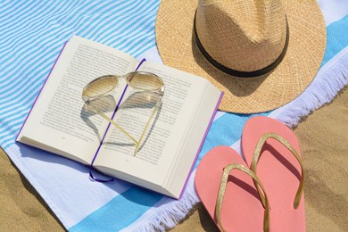 Photo of Beach towel with book, straw hat, sunglasses and flip flops on sand