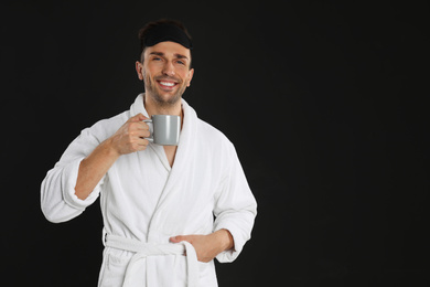 Young man in bathrobe with cup of coffee on black background. Space for text