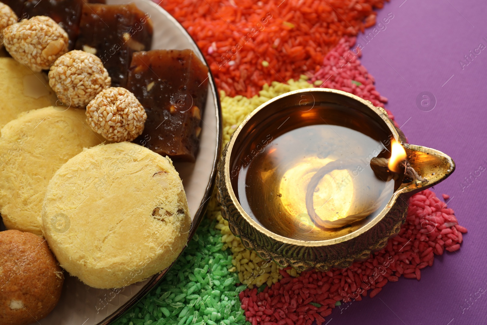 Photo of Diwali celebration. Tasty Indian sweets, diya lamp and colorful rangoli on violet table, above view