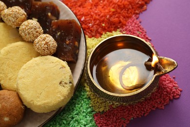 Photo of Diwali celebration. Tasty Indian sweets, diya lamp and colorful rangoli on violet table, above view