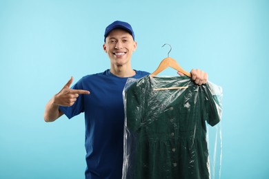 Dry-cleaning delivery. Happy courier holding dress in plastic bag on light blue background