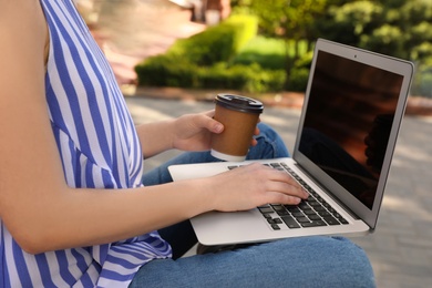 Image of Young woman with paper cup of coffee working on laptop outdoors, closeup