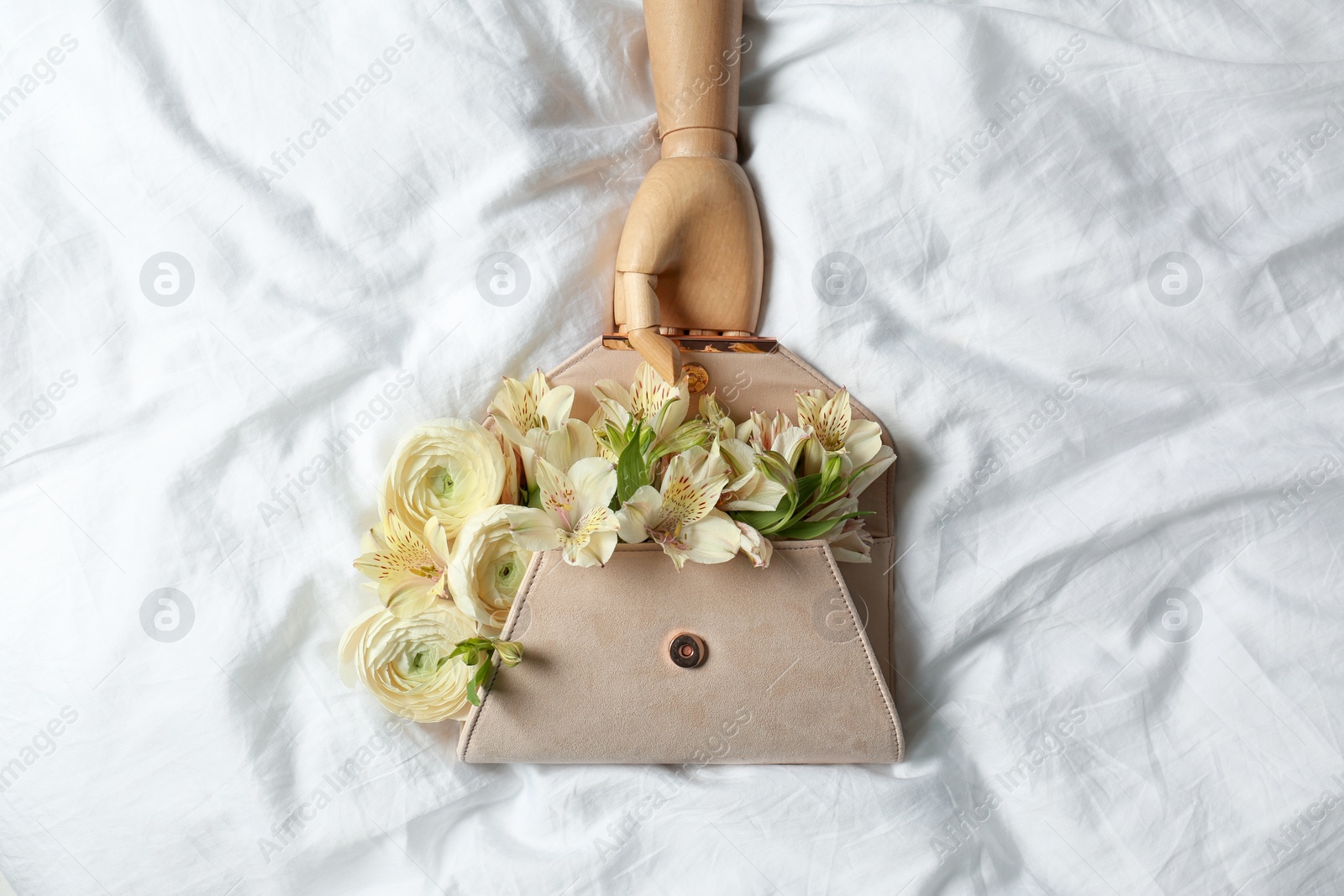 Photo of Stylish clutch with spring flowers and wooden mannequin hand on crumpled sheet, flat lay