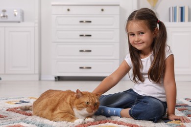 Smiling little girl petting cute ginger cat on carpet at home, space for text