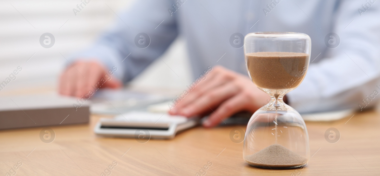 Photo of Hourglass with flowing sand on desk. Man using calculator indoors, selective focus