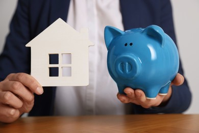 Photo of Woman holding piggy bank and house model at wooden table, closeup