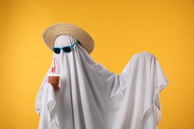 Photo of Person in ghost costume, sunglasses and straw hat holding glass of drink on yellow background