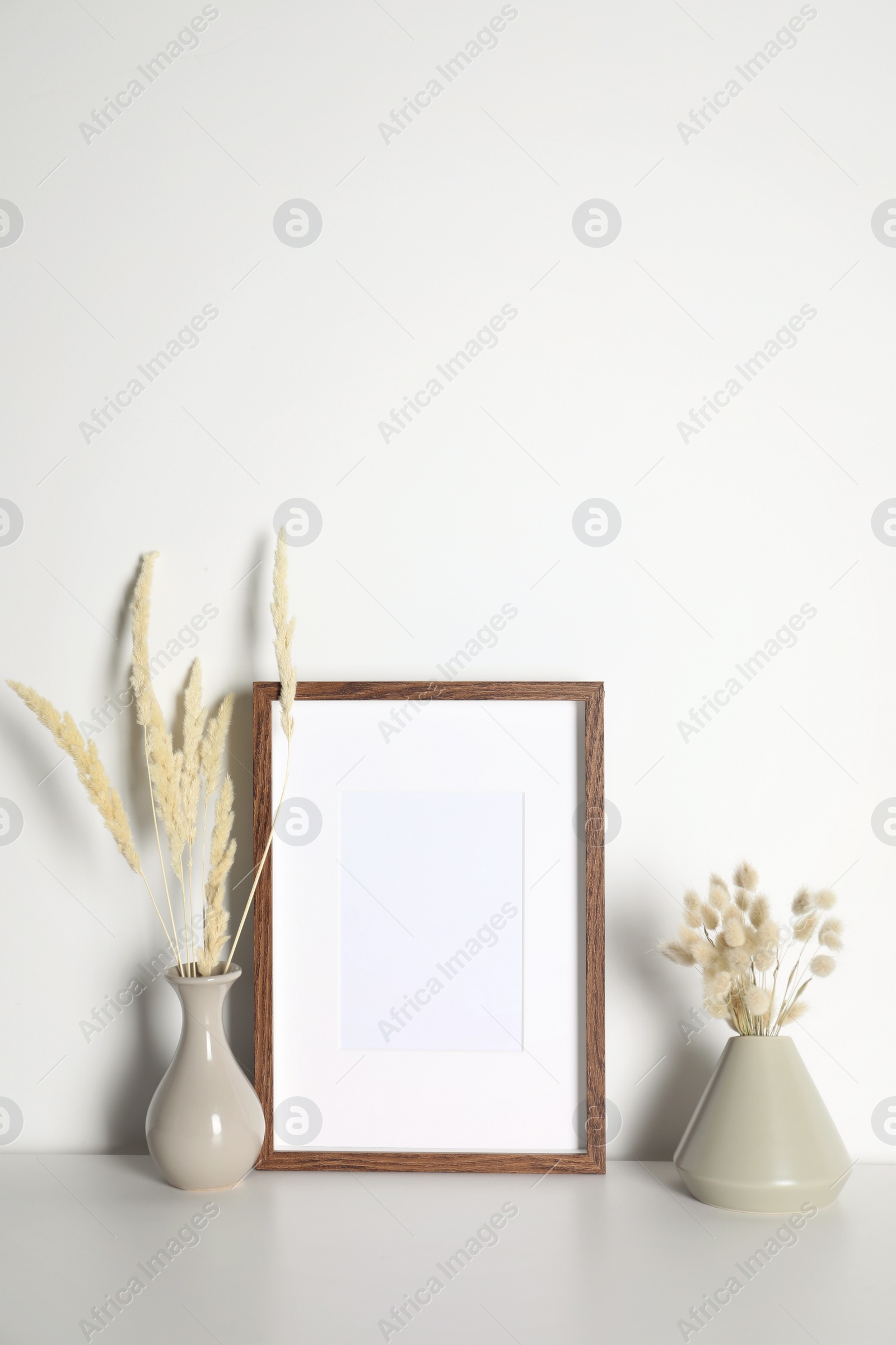 Photo of Empty photo frame and vases with dry decorative spikes on white table. Mockup for design