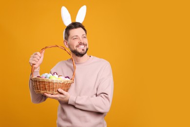 Happy man in bunny ears headband holding wicker basket with painted Easter eggs on orange background. Space for text
