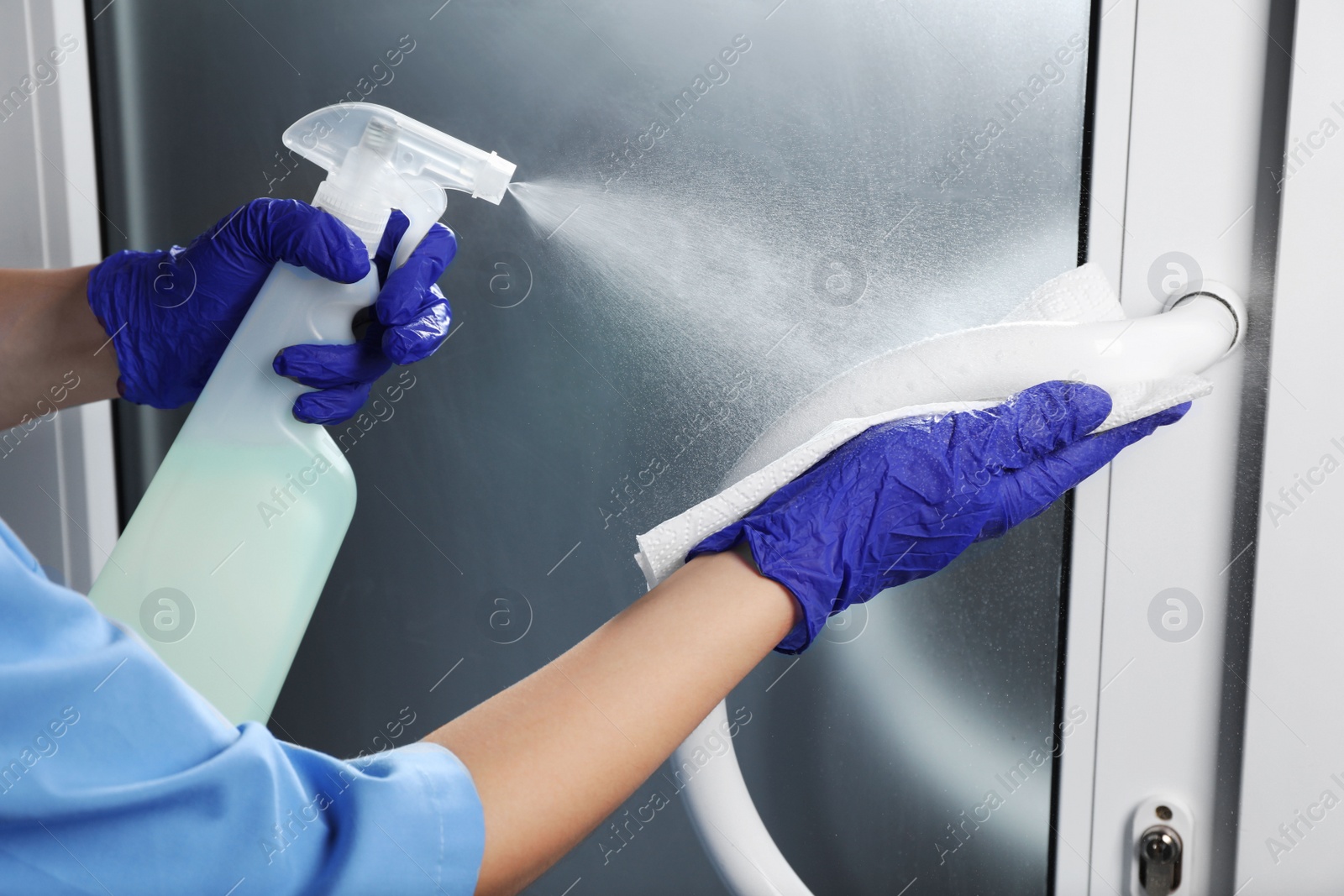 Photo of Woman in gloves cleaning door handle with paper towel and detergent indoors, closeup