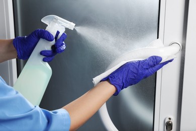 Photo of Woman in gloves cleaning door handle with paper towel and detergent indoors, closeup