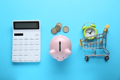 Photo of Calculator, piggy bank, coins and alarm with cart on light blue background, flat lay