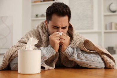 Sick man wrapped in blanket with tissue blowing nose at wooden table indoors. Cold symptoms