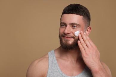 Photo of Handsome man applying cream onto his face on pale brown background, space for text