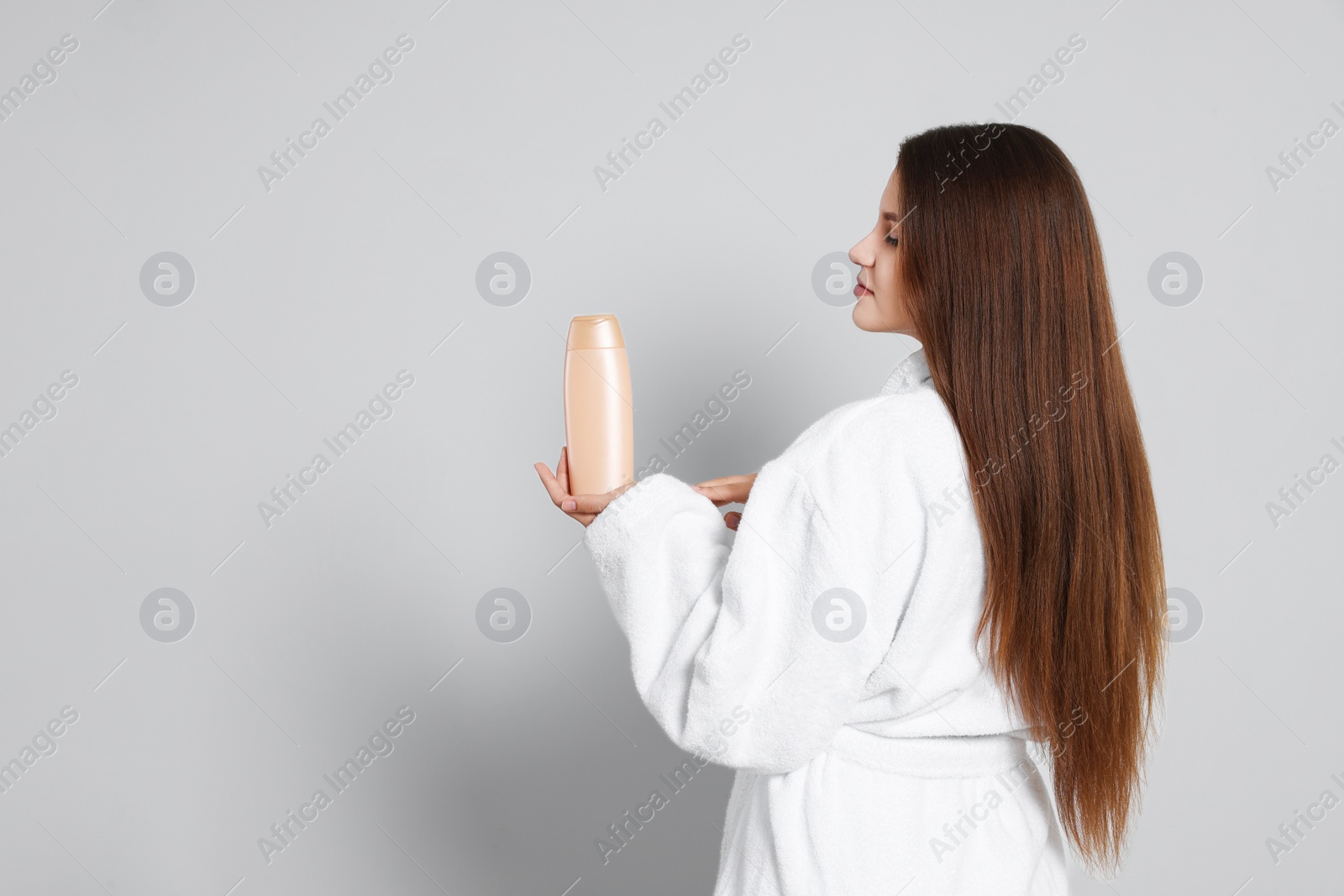 Photo of Beautiful young woman in bathrobe holding bottle of shampoo on light grey background. Space for text