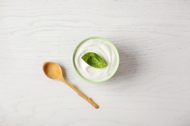 Photo of Bowl of fresh sour cream with basil and spoon on white wooden table, flat lay