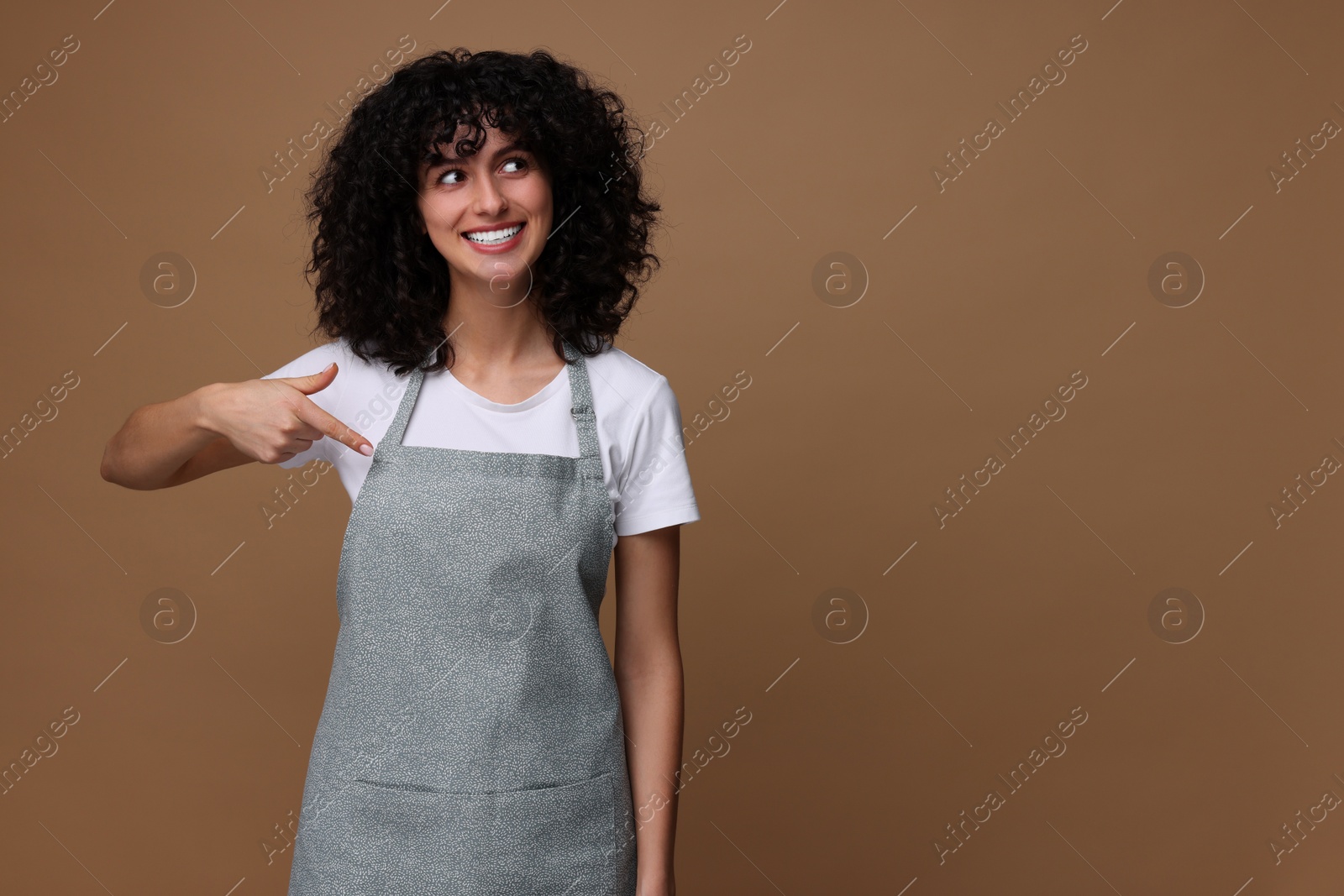 Photo of Happy woman pointing at kitchen apron on brown background, space for text. Mockup for design