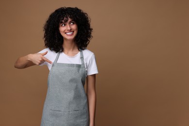 Happy woman pointing at kitchen apron on brown background, space for text. Mockup for design